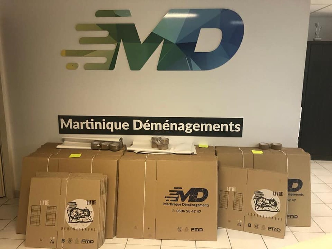 Des cartons de déménagement et des rouleaux de ruban adhésif sont empilés au sol devant un mur portant le logo et le nom de Martinique Déménagements.