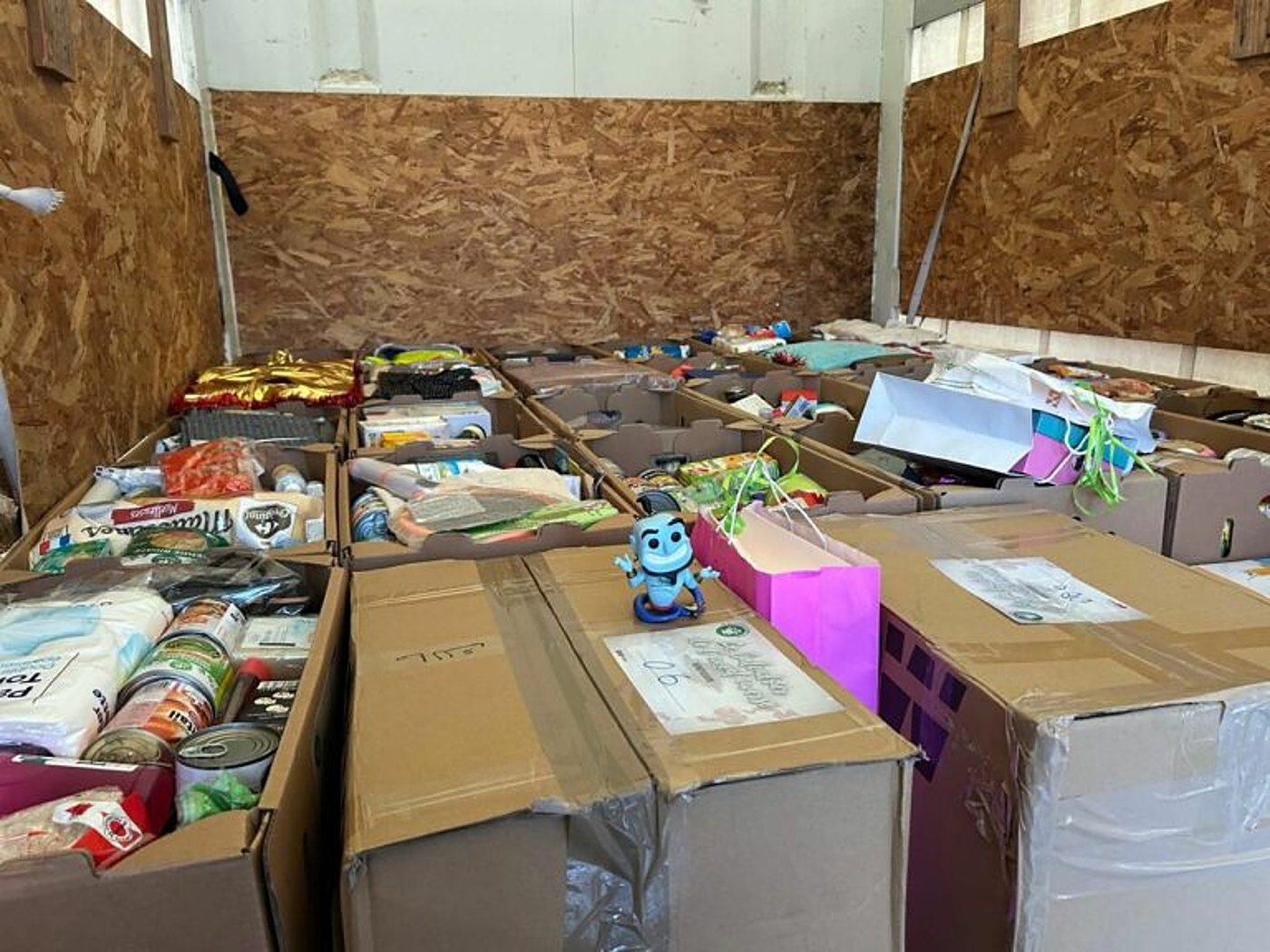 Intérieur d'un camion aux parois en bois aggloméré rempli de cartons de conserves, de denrées et de sacs cadeaux, avec une petite figurine bleue posée sur l'un des cartons.