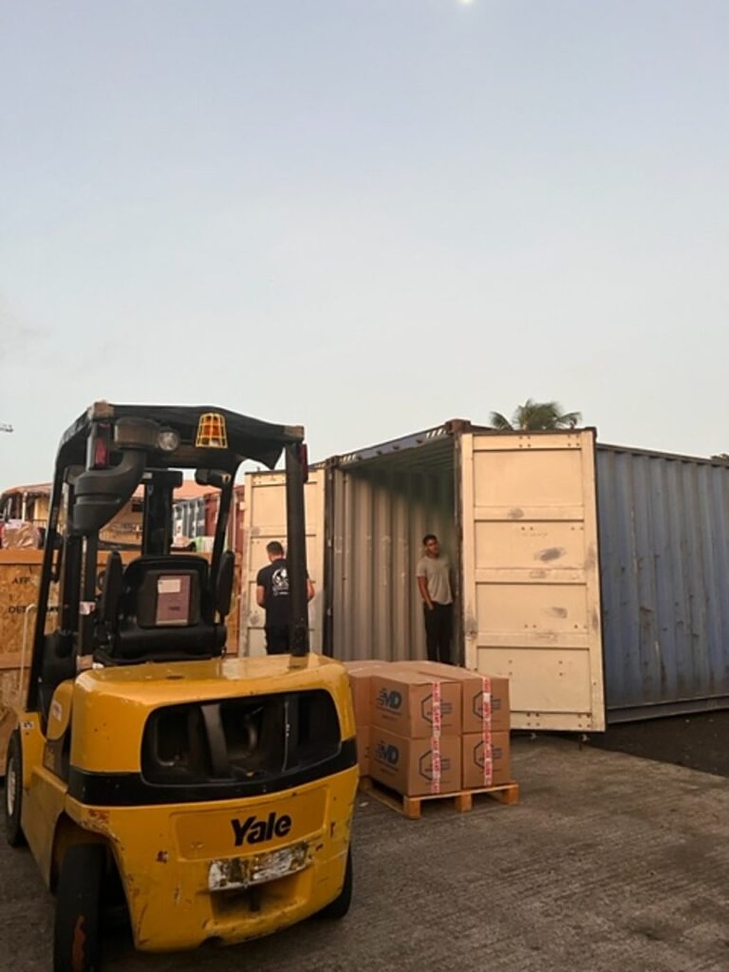 Un chariot élévateur jaune Yale est garé près d'un conteneur ouvert et d'une palette de cartons, avec deux hommes à l'arrière-plan au crépuscule.