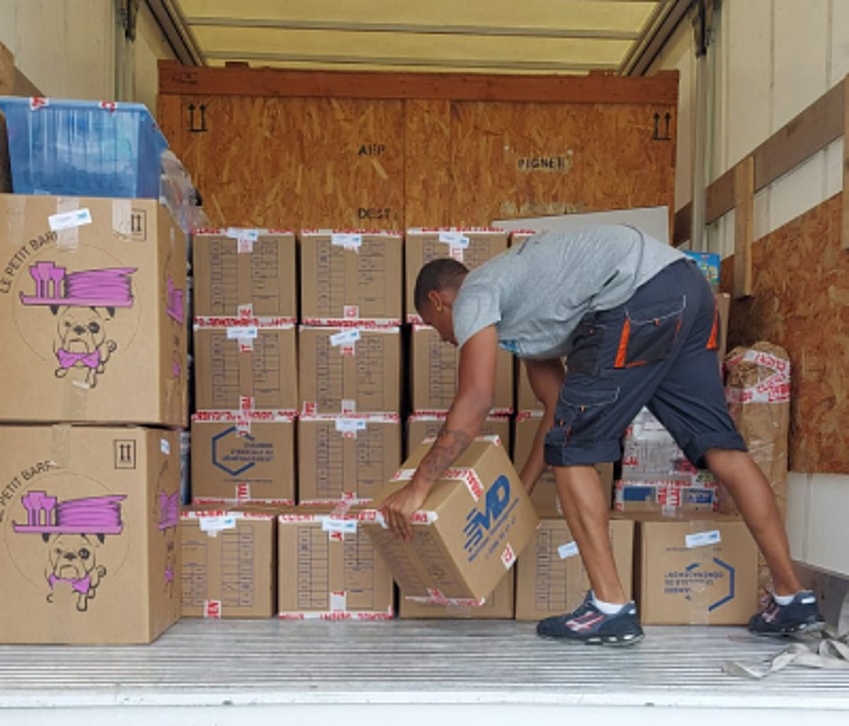 Un déménageur en t-shirt gris et short sombre soulève un carton pour l'empiler avec d'autres cartons dans la remorque d'un camion.