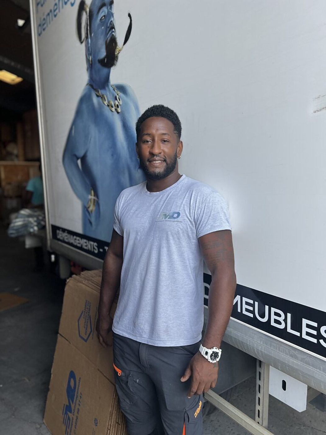 Un déménageur en t-shirt gris se tient souriant devant un camion blanc décoré d'un grand personnage bleu, à côté de cartons de déménagement.