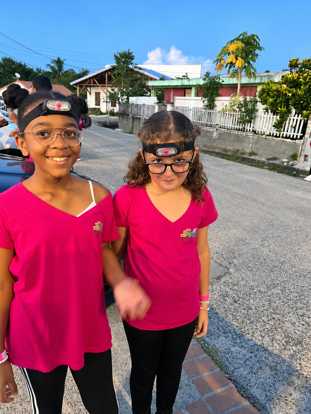 Deux jeunes filles souriantes portant des t-shirts roses et des lampes frontales posent côte à côte dans une rue au crépuscule, devant des maisons et un papayer.