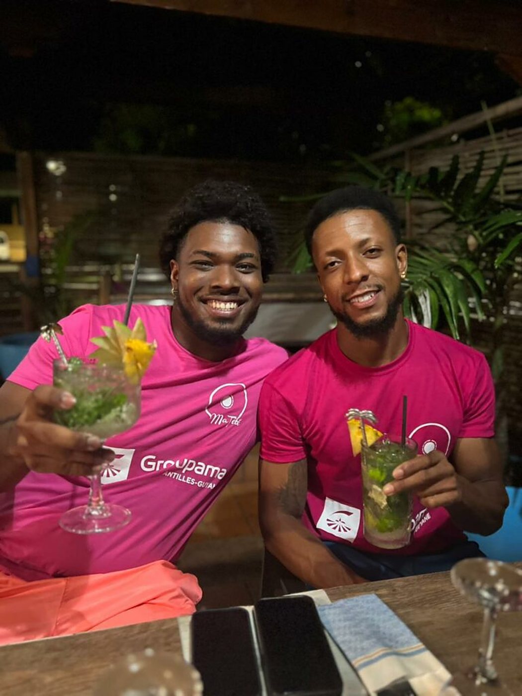Deux hommes souriants portant des t-shirts roses Groupama tiennent chacun un mojito à une table en bois, le soir, sur une terrasse entourée de plantes.