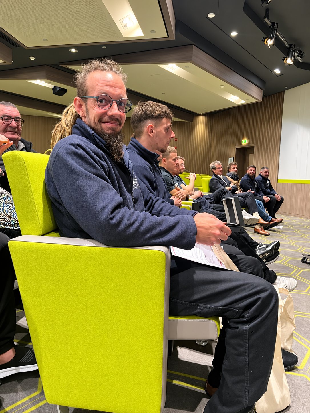 Un homme barbu à lunettes et dreadlocks sourit à l'appareil, assis sur un fauteuil vert anis, entouré de collègues en vestes sombres dans une salle de conférence.