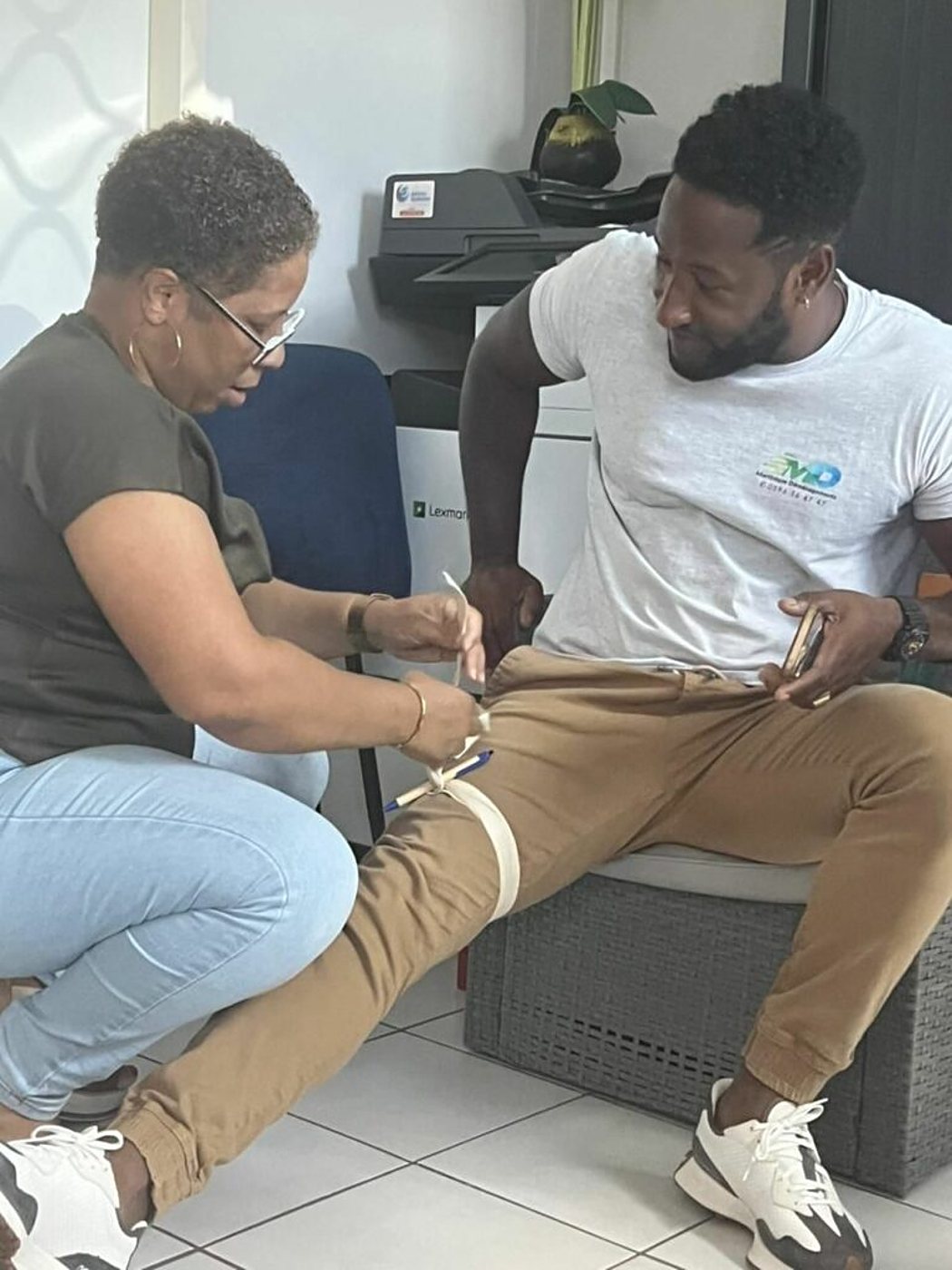 Dans un bureau, une femme à lunettes applique un garrot sur la cuisse d'un homme assis sur un pouf, qui regarde son téléphone.