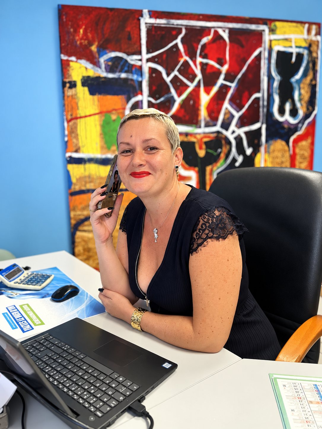 Une femme aux cheveux courts et blonds sourit en téléphonant, assise à un bureau avec un ordinateur portable, devant un grand tableau coloré sur un mur bleu.