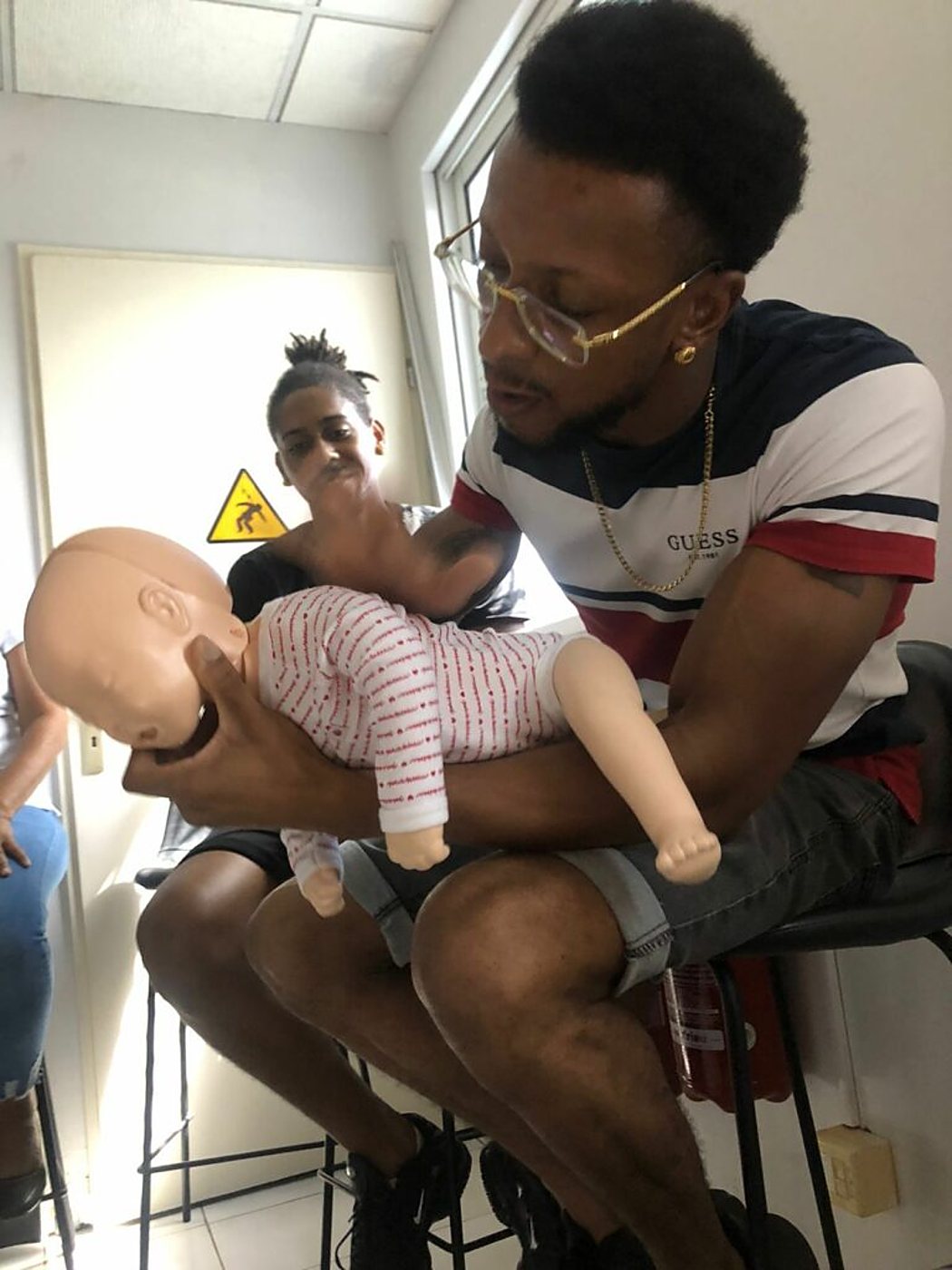 Un homme à lunettes tient un mannequin de nourrisson sur son avant-bras pour démontrer un geste de secours devant d'autres participants assis dans une salle de formation.