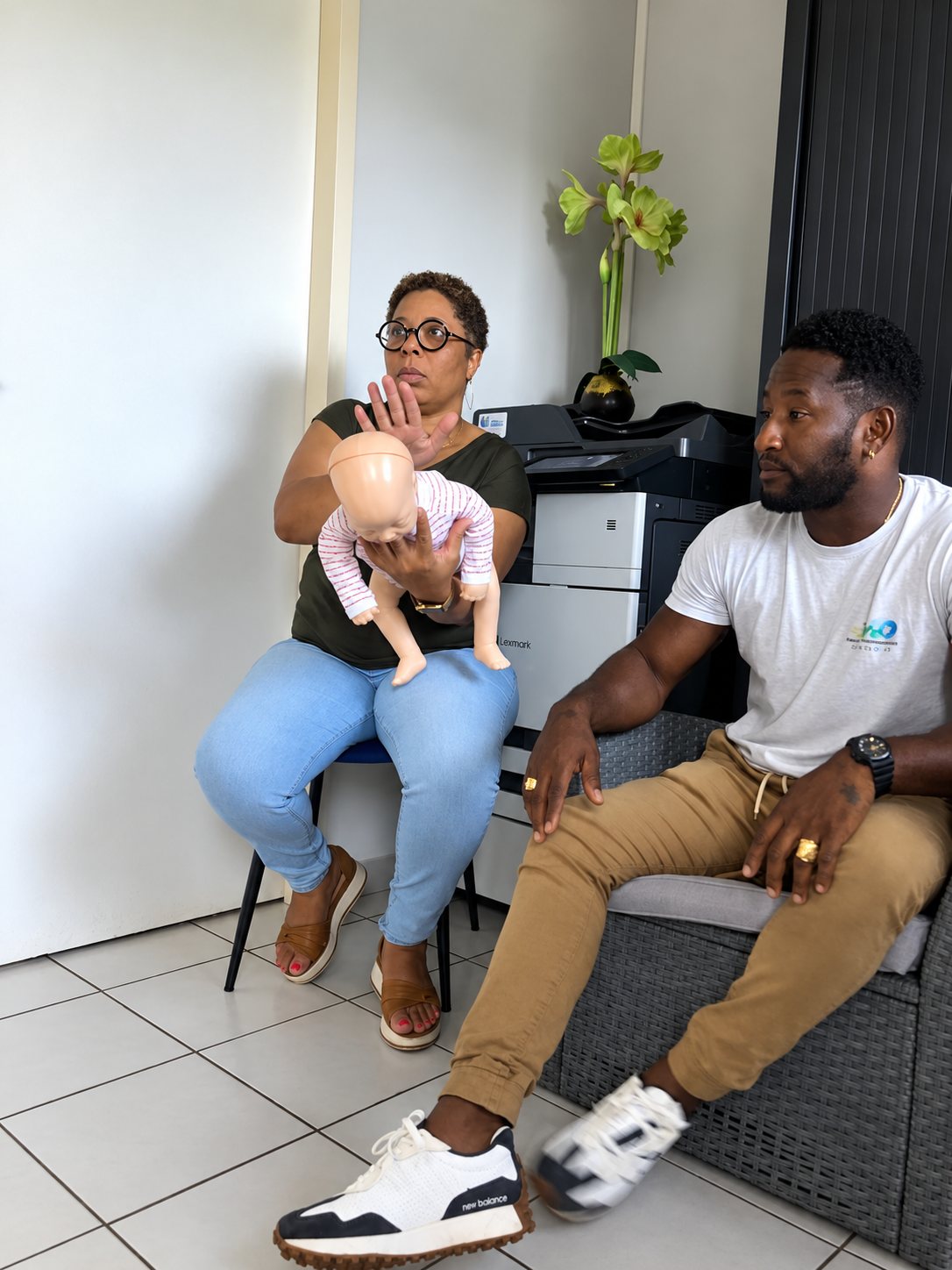 Une formatrice assise tient un mannequin de nourrisson pour montrer un geste de secours, à côté d'un homme assis sur un fauteuil en résine tressée, dans un bureau avec une imprimante.