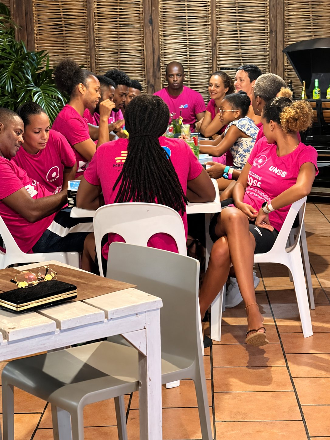 Un groupe d'adultes et d'enfants vêtus de t-shirts roses est assis autour d'une longue table dans un restaurant aux murs de bambou tressé.