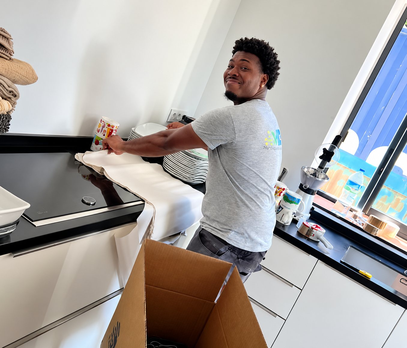 Un homme en t-shirt gris sourit en déballant de la vaisselle posée sur un plan de travail noir dans une cuisine blanche, à côté d'un carton ouvert.