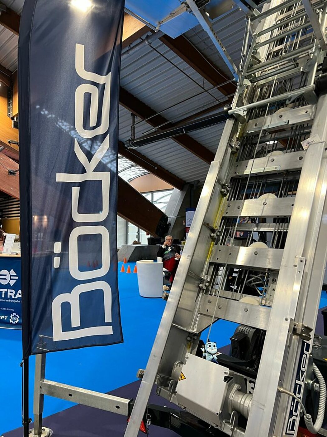 Un monte-matériaux en aluminium de la marque Böcker est exposé sur un stand de salon, à côté d'une oriflamme bleue portant le nom Böcker, sous une charpente en bois.