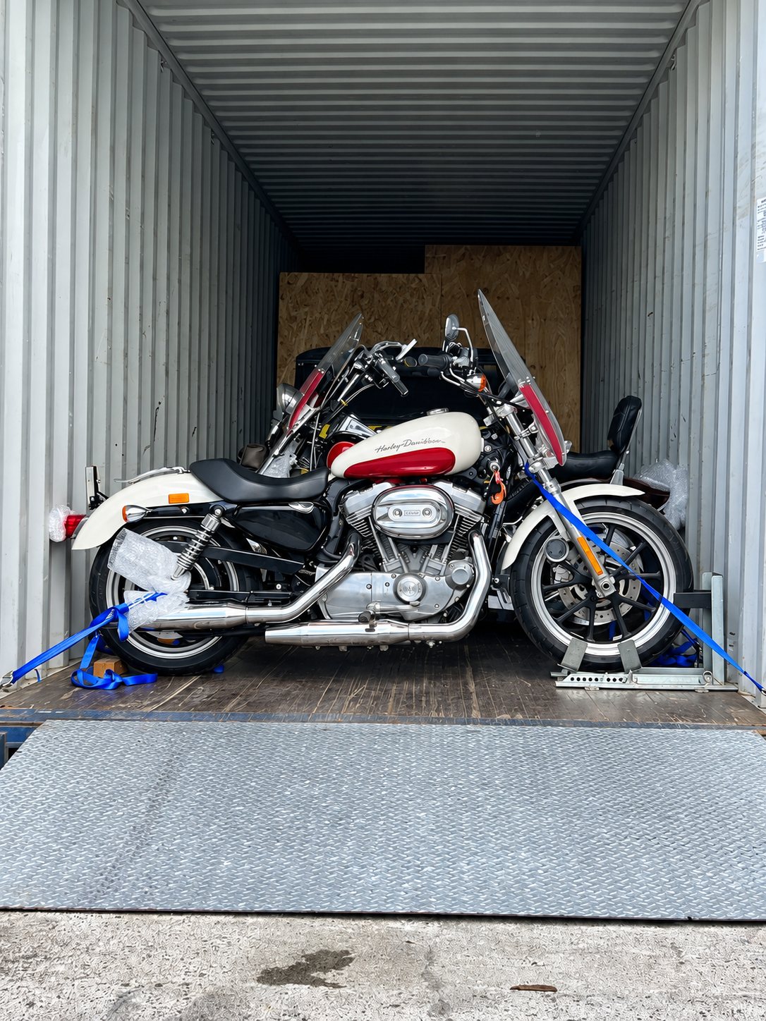Une moto Harley-Davidson crème et rouge est fixée par des sangles bleues à l'intérieur d'un conteneur, avec une autre moto derrière elle.
