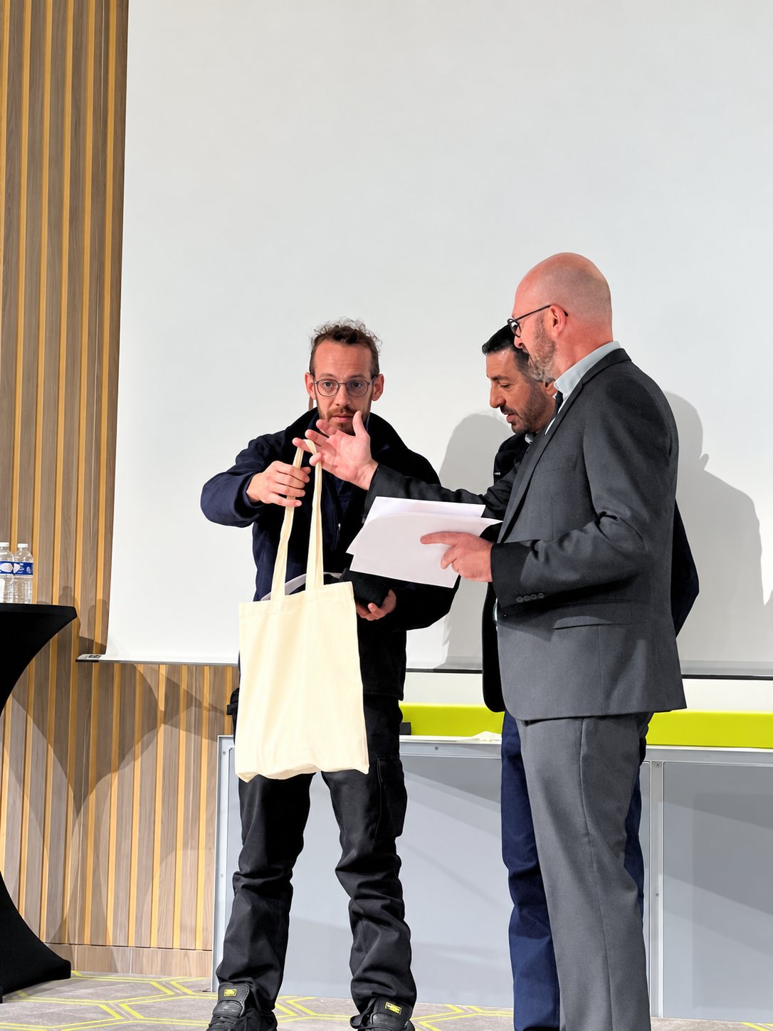 Trois hommes sur une scène lors d'une remise : l'un tient un tote bag en toile écrue tandis qu'un autre tient des feuilles.