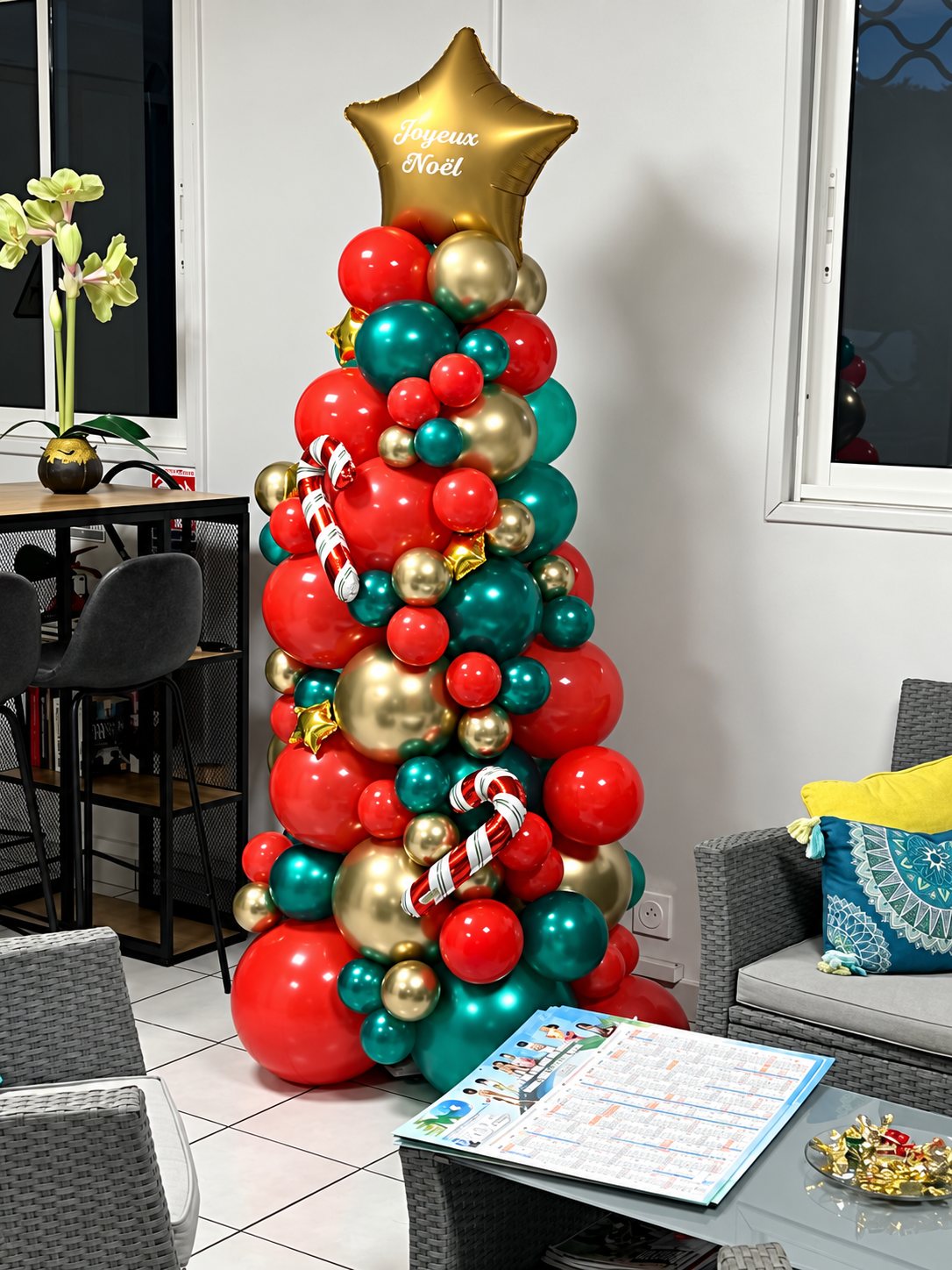 Un sapin de Noël réalisé en ballons rouges, verts et dorés, surmonté d'une étoile dorée marquée « Joyeux Noël », installé dans un salon près d'une table basse.